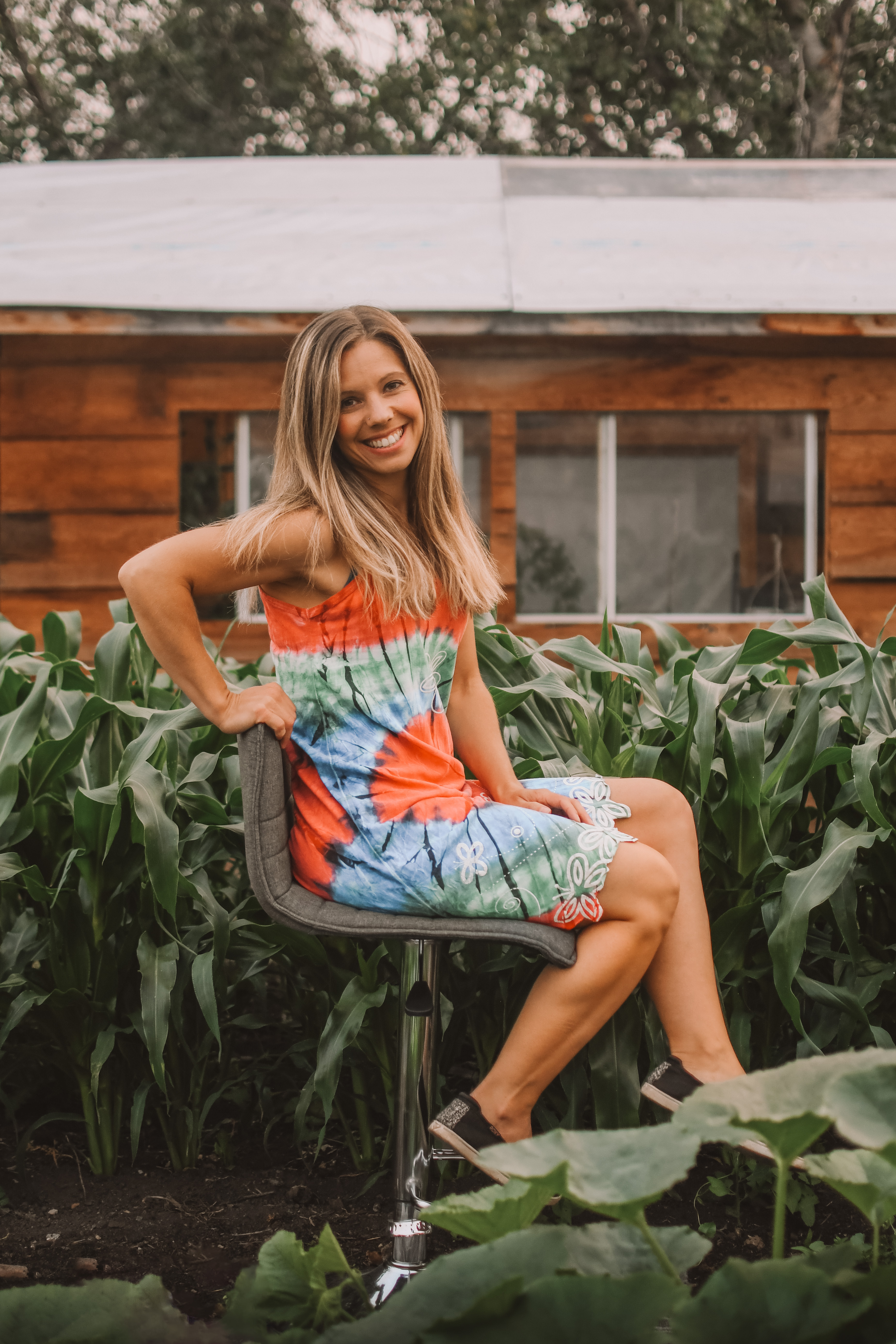 Amy Soudek smiling, seated in front of tall corn plants and a wooden cabin in her garden.